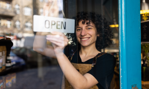 A woman standing behind a glass door flipping a sign that reads open