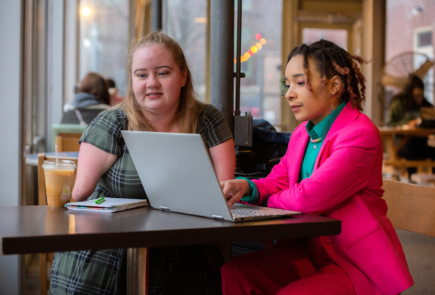 Two women sitting at a table looking at a computer in a coffee shop 